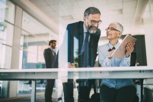 A retired couple reviews financial papers at a kitchen table with a notebook and calculator.